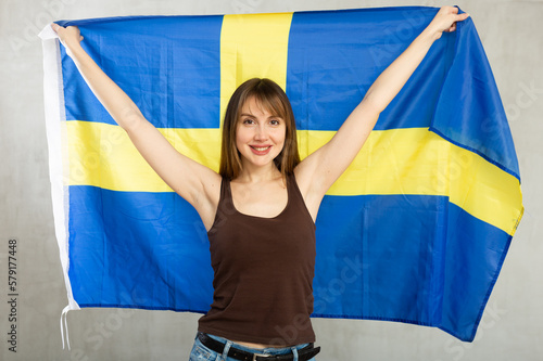 Portrait of smiling young woman holding in hands national flag of Sweden  posing against gray studio background