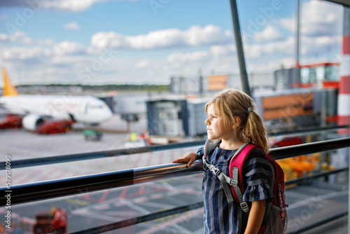 Little girl at the airport waiting for boarding at the big window. Cute kid stands at the window against the backdrop of airplanes. Looking forward to leaving for a family summer vacation