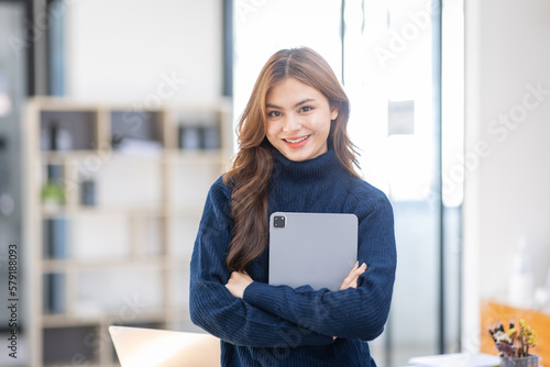 portrait of young asian woman, company worker at workplace, smiling and holding digital tablet, standing over office background