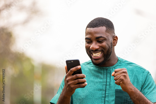 Excited Young African Man Holding smartphone outdoors, happy to win lottery. photo