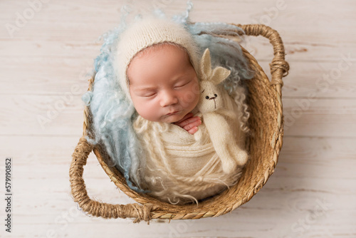 Top view of a newborn baby girl sleeping in a white cape with a blue bandage and a flower on her head, in wicker basket with blue fur and a rabbit. Beautiful portrait of a little girl 7 days, one week
