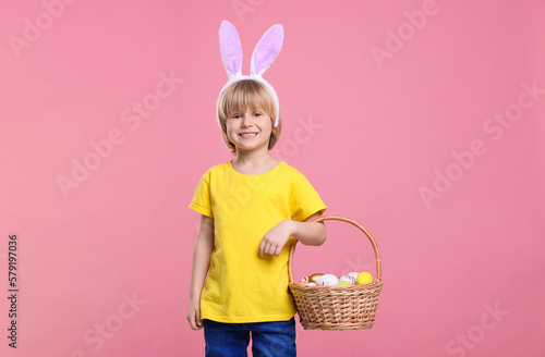 Happy boy in bunny ears headband holding wicker basket with painted Easter eggs on pink background