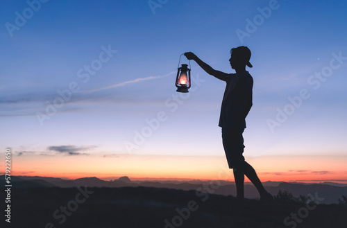 Silhouette of a man holding a lit kerosene lamp to light his way in the dark, with sky after sunset in the background. Concept of faith and hope in the face of adversity.
