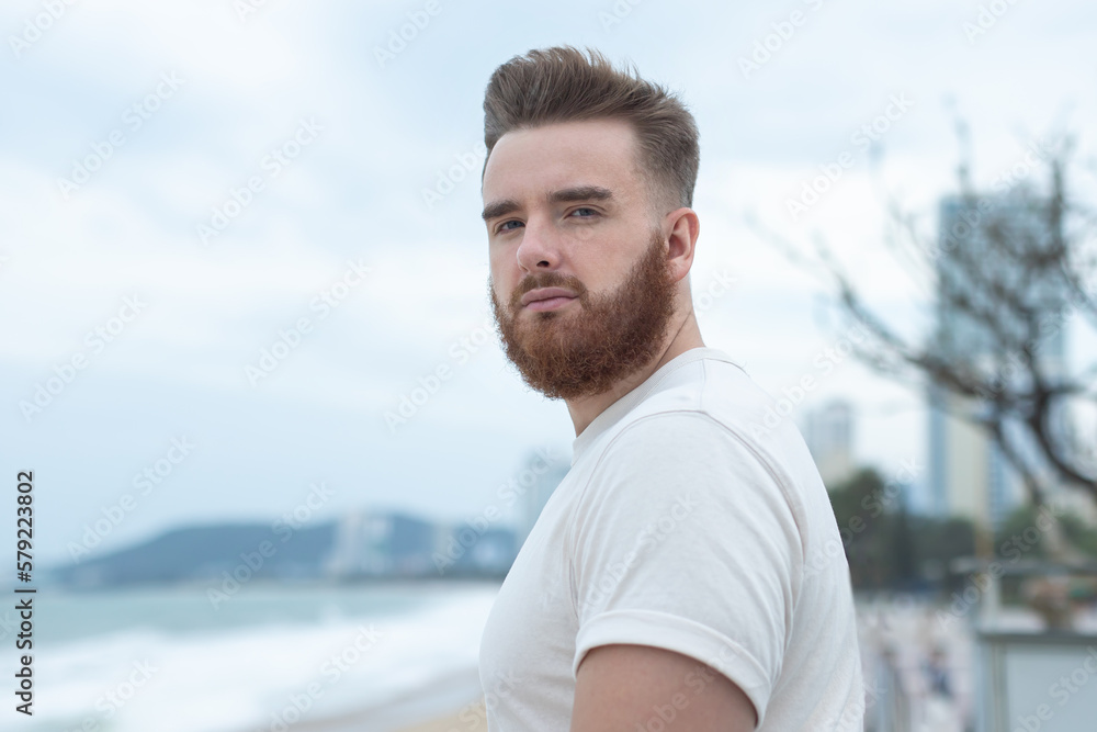 Fototapeta premium Portrait of young handsome bearded pensive man with beard is enjoy walk on summer beach near sea, breathe deep fresh air in tropical country. Summer vacation.