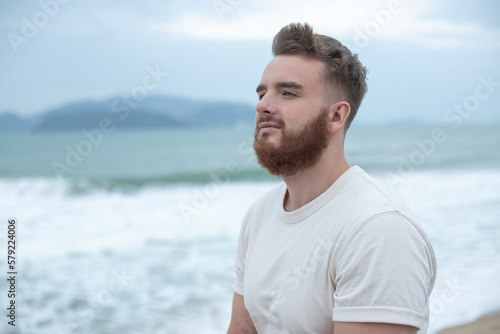 Portrait of young handsome bearded pensive man with beard is enjoy walk on summer beach near sea, breathe deep fresh air in tropical country. Summer vacation.