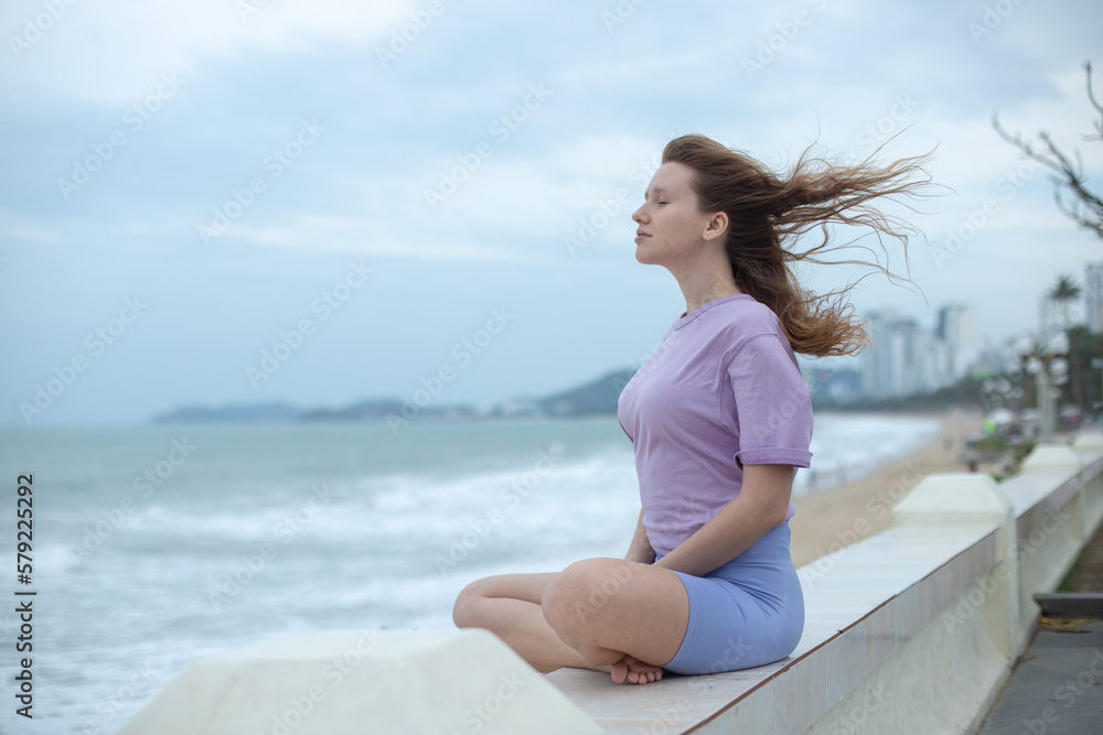 Young calm sad serious woman is sitting on embankment near sea, ocean on beach, girl thinking, meditate on natural background 