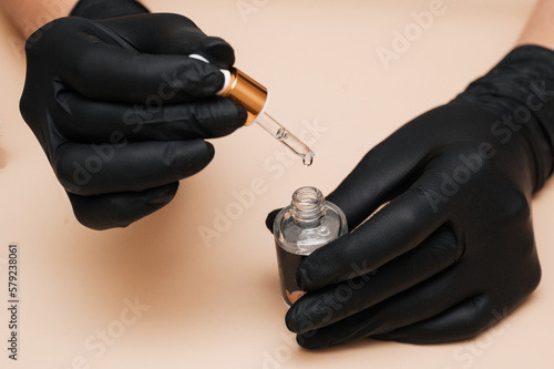 Close-up. The manicure master holds a tube of nail polish on a light background in his gloved hands.