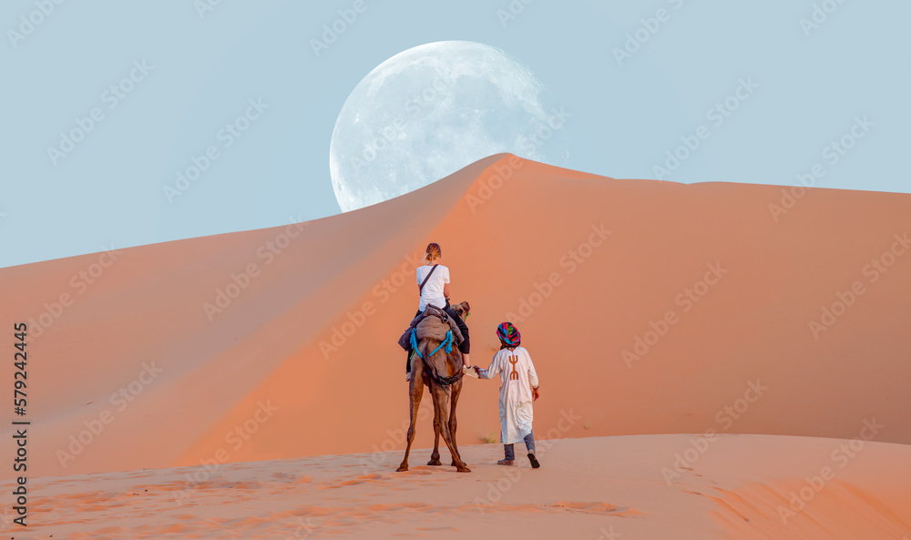 A woman riding a camel across the thin sand dunes of the in Western Sahara Desert with full moon - Morocco, Africa - obrazy, fototapety, plakaty 