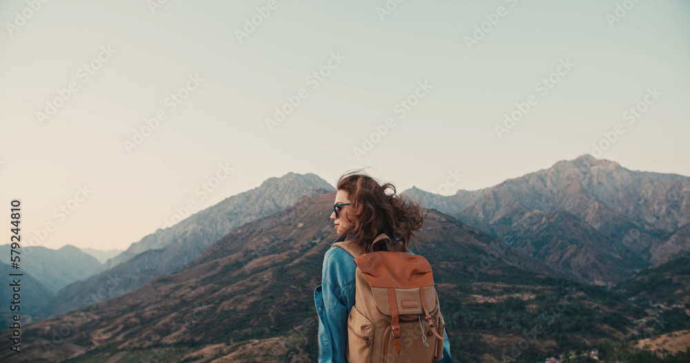 Happy slim girl is standing at sunset mountains and enjoying the view. Hair blowing in the wind.Girl admiring mountain landscape. A woman standing alone in sunset scene