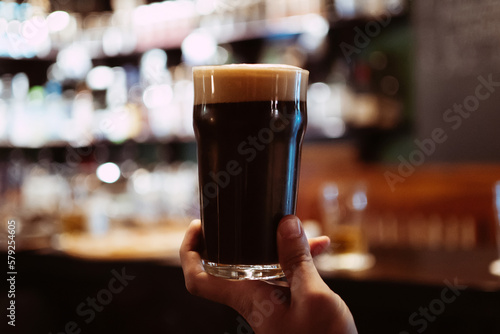 Fotografia glass of dark stout beer with foam and bubbles in hand of a man in pub behind th