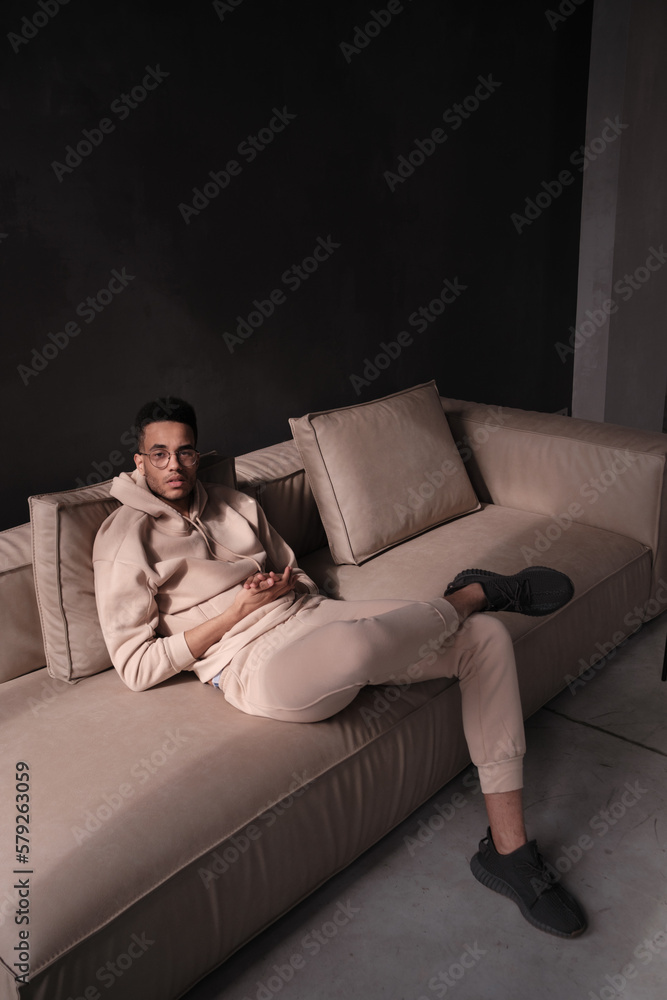 Obraz premium Portrait of a cheerful young african american man 20s wearing casual hoody and glasses sitting on sofa against black background. copy space isolated