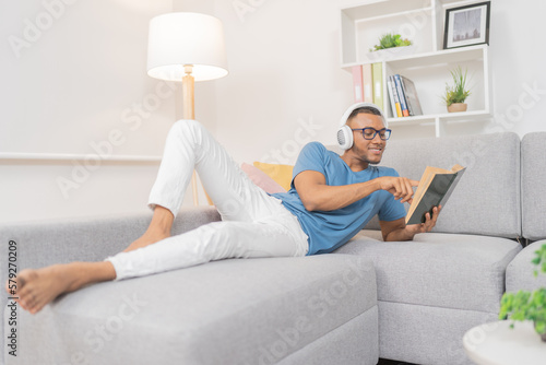 Young man, smiling and relaxing, listening to music with his headphones while reading a book after a long day at work