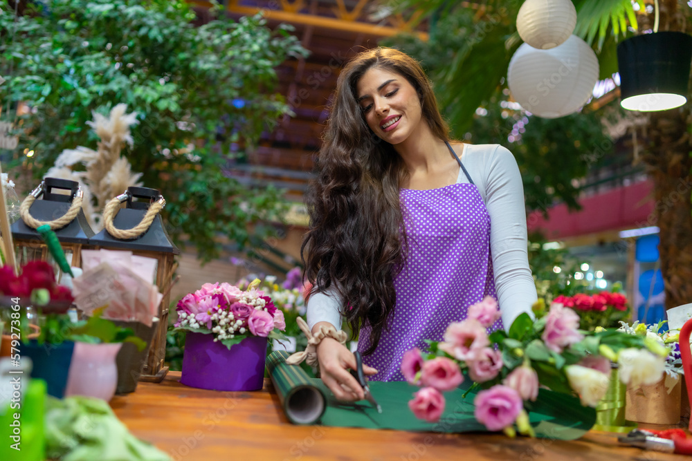 Fototapeta premium A beautiful florist stands behind a counter in a flower shop and makes a bouquet using garden scissors and decorative paper
