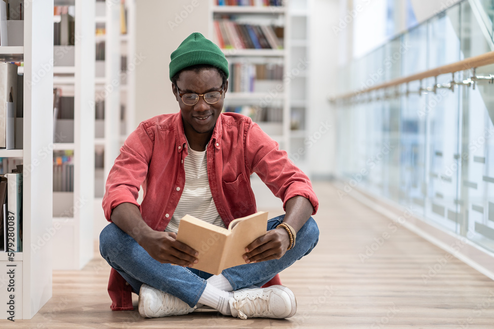 Interested Black student man sitting on floor in library. Focused African American guy preparing materials for upcoming lecture reading book in university. Higher education, bibliophile concept.