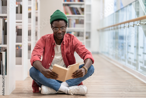 Interested Black student man sitting on floor in library. Focused African American guy preparing materials for upcoming lecture reading book in university. Higher education, bibliophile concept.