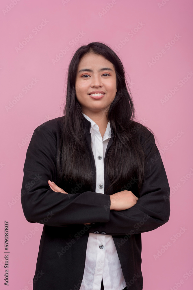 A young southeast asian female employee in a black jacket with arms crossed. Isolated on a light pink background. A young southeast asian female employee in a black jacket with arms crossed. Isolated on a light pink background.