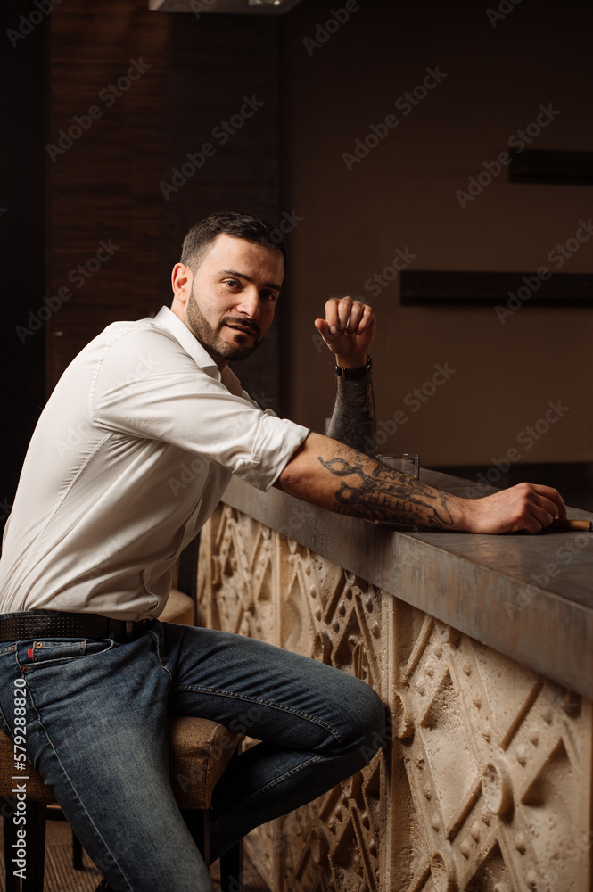 Handsome bearded male in shirt and jeans with glass of whiskey and cigar in the bar or pub Handsome bearded male in shirt and jeans with glass of whiskey and cigar in the bar or pub
