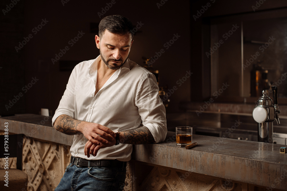 Handsome bearded male in shirt and jeans with glass of whiskey and cigar in the bar or pub Handsome bearded male in shirt and jeans with glass of whiskey and cigar in the bar or pub