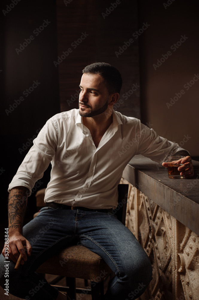 Handsome bearded male in shirt and jeans with glass of whiskey and cigar in the bar or pub