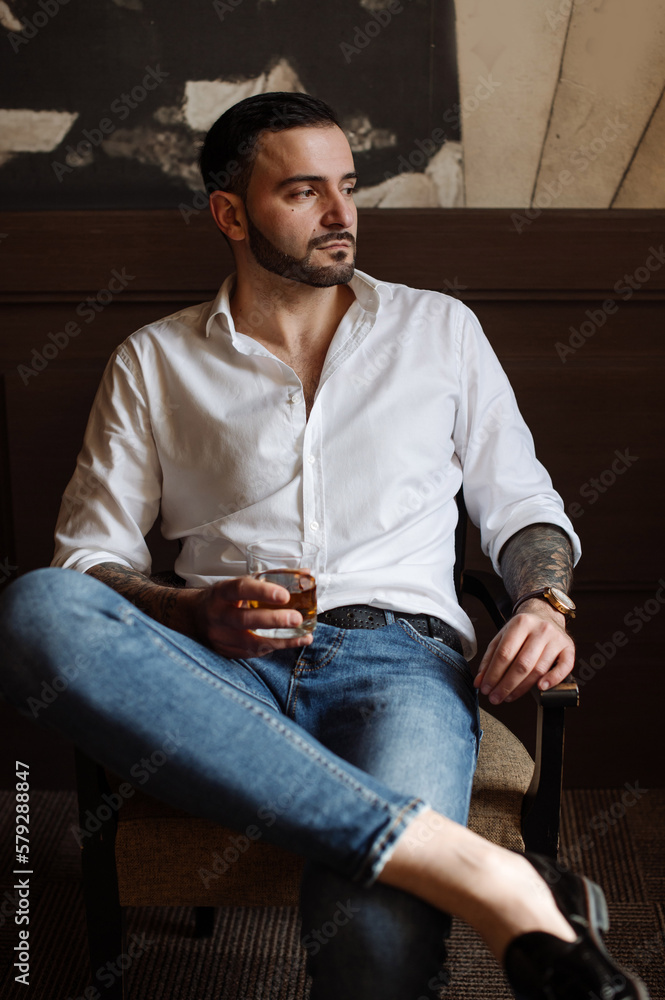 Handsome bearded male dressed in white shirt and jeans is sitting on chair indoor the room Handsome bearded male dressed in white shirt and jeans is sitting on chair indoor the room