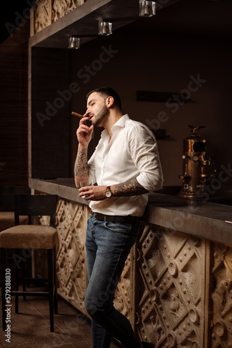 Handsome bearded male in shirt and jeans with glass of whiskey and cigar in the bar or pub