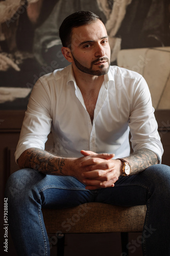 Handsome bearded male dressed in white shirt and jeans is sitting on chair indoor the room