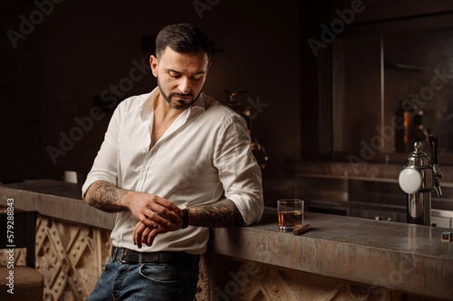 Handsome bearded male in shirt and jeans with glass of whiskey and cigar in the bar or pub
