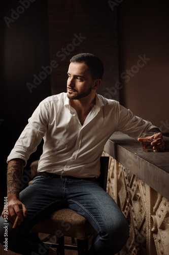 Handsome bearded male in shirt and jeans with glass of whiskey and cigar in the bar or pub
