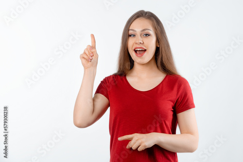 Surprised lovely girl points finger up, shows advertisement on blank white space. Young brunette female looking amazed by promotion offer, stands over white background