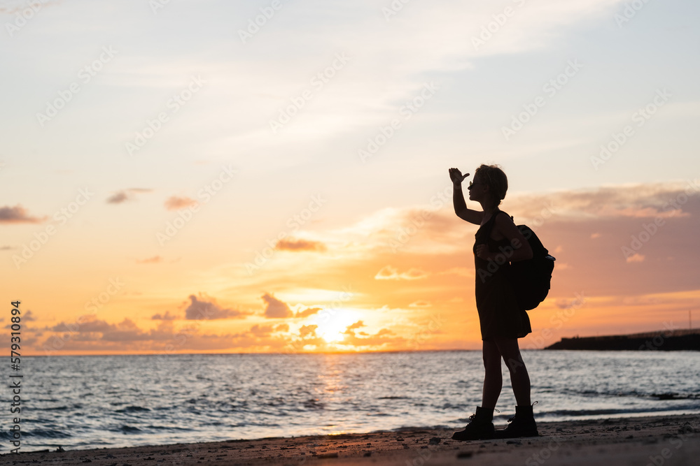 Silhouette of a traveler girl with a backpack, standing by the sea on the beach, meeting the sunset.