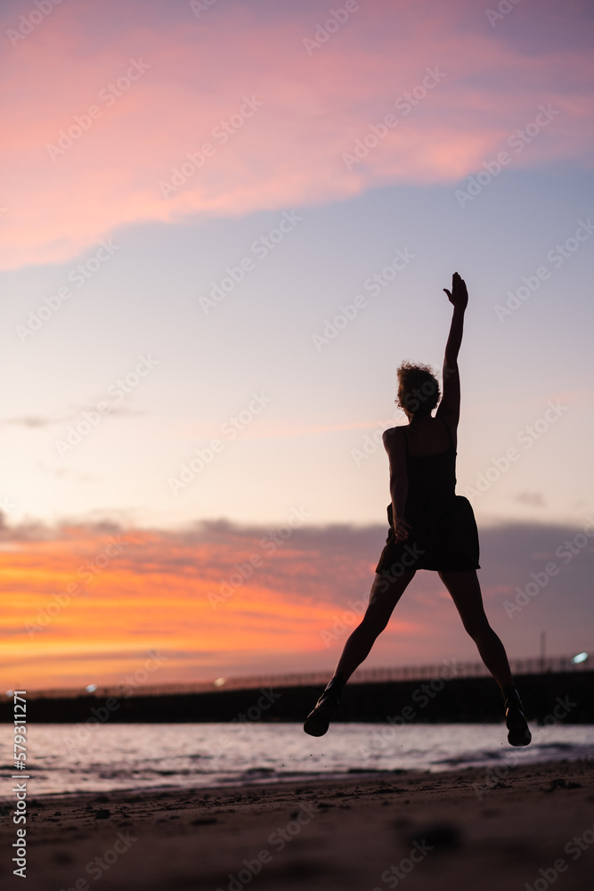 A young girl admires the sunset on the ocean, enjoys life, arms outstretched to the sides, rear view.