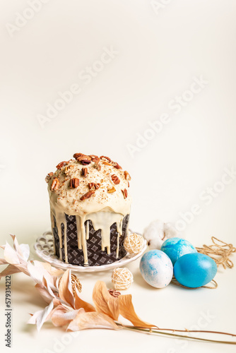 Traditional orthodox Easter cake - Paska with white chocolate and nuts on white round plate and blue colored Easter eggs on light beige background in rustic style.