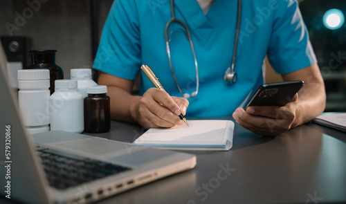 male medicine doctor, physician or practitioner in lab room writing on blank notebook and work on laptop computer with medical stethoscope on the desk at hospital. Medic tech concept.