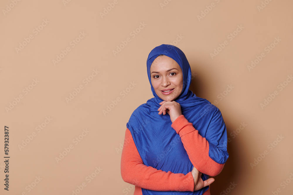 Confident middle-aged elegant charming Arab Muslim woman, dressed in blue hijab and orange dress, looking at camera, isolated beige background with copy space. Islamic fashion. Middle-Eastern culture Confident middle-aged elegant charming Arab Muslim woman, dressed in blue hijab and orange dress, looking at camera, isolated beige background with copy space. Islamic fashion. Middle-Eastern culture