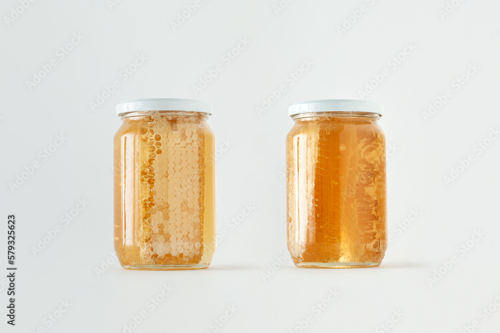 Glass jars with honeycomb and golden honey on isolated pastel white background. Minimal creative idea of alternative therapy or eating raw, healthy, natural sweet food. Beekeeping products concept. Glass jars with honeycomb and golden honey on isolated pastel white background. Minimal creative idea of alternative therapy or eating raw, healthy, natural sweet food. Beekeeping products concept.