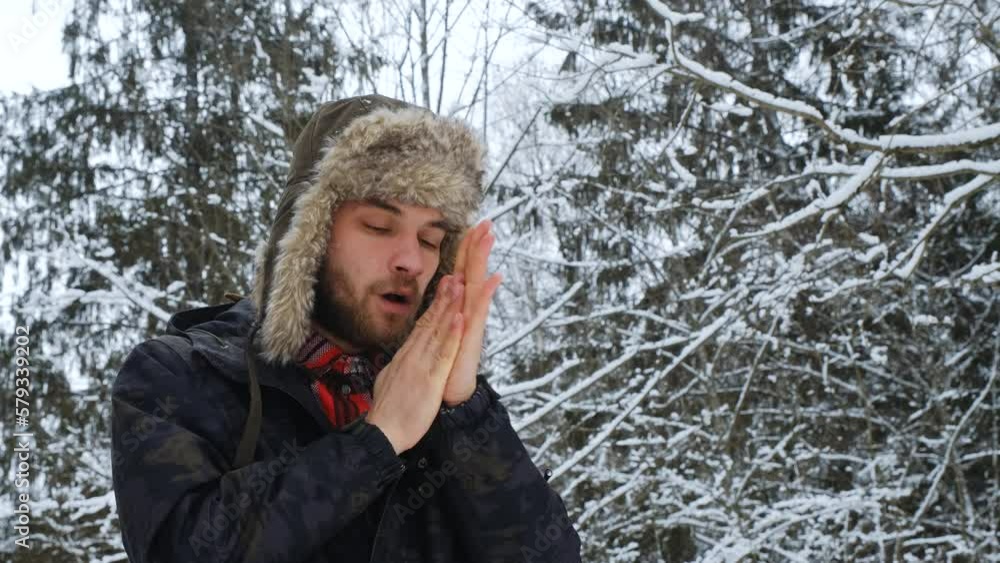 Man froze on walk outside in winter and trying to warm up, warming hands with breath. Concept of problem and peculiarities nature of North. Guy in hat with earflaps in snow park. Man froze on walk outside in winter and trying to warm up, warming hands with breath. Concept of problem and peculiarities nature of North. Guy in hat with earflaps in snow park.