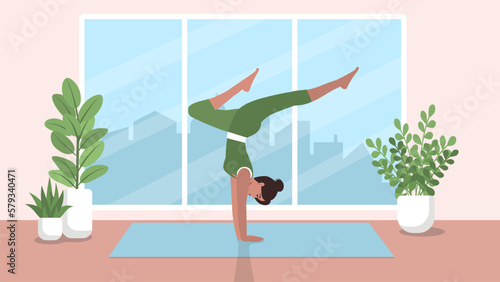 An athletic woman performs a handstand of yoga or gymnastics in a room with flowers and a large window overlooking the city. The concept of a healthy and active lifestyle, sports.