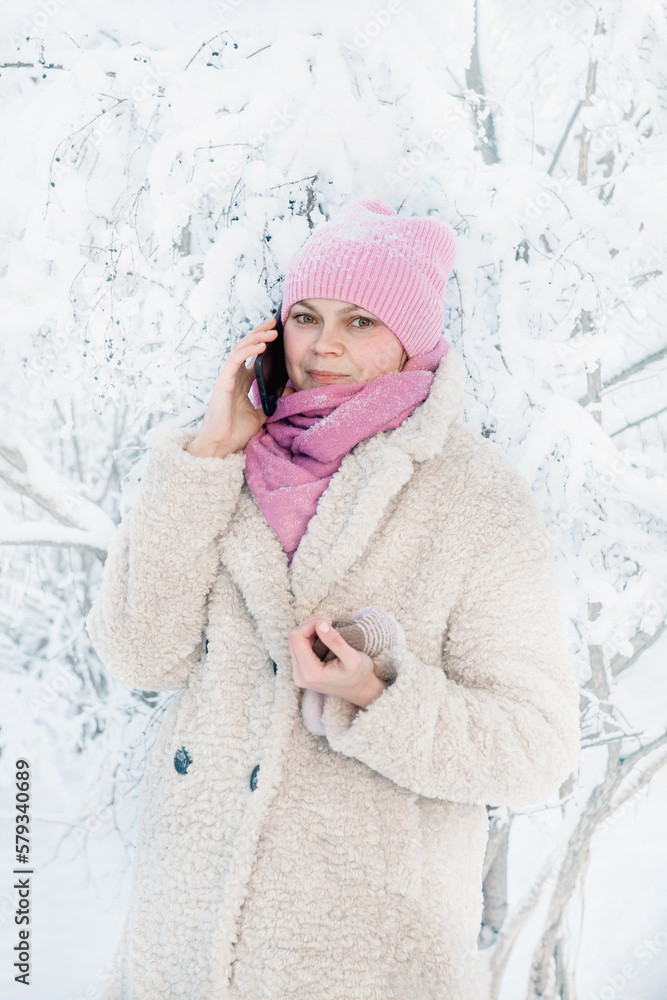 A young woman in a warm hat and scarf. The concept of winter fashion. A happy woman is talking on the phone on a snowy winter day. A woman smiles against the background of snowy winter nature A young woman in a warm hat and scarf. The concept of winter fashion. A happy woman is talking on the phone on a snowy winter day. A woman smiles against the background of snowy winter nature