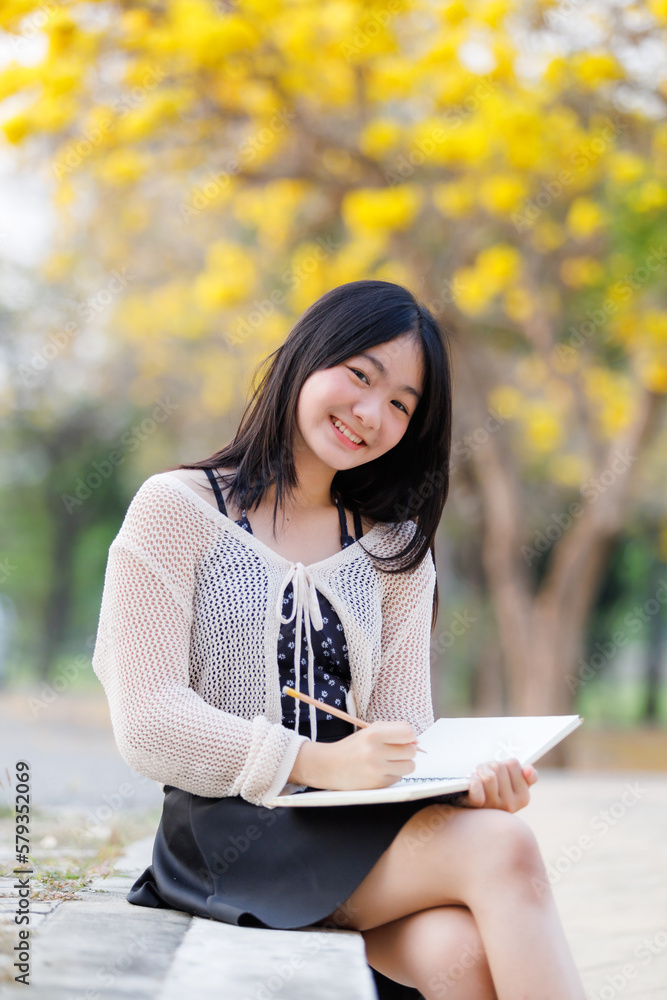 Portrait cute smiles Asian of attractive young teenage girl a holding diary book writing note at Yellow Tabebuia Chrysotricha flowers with the park in spring day at Evening background in Thailand. Portrait cute smiles Asian of attractive young teenage girl a holding diary book writing note at Yellow Tabebuia Chrysotricha flowers with the park in spring day at Evening background in Thailand.