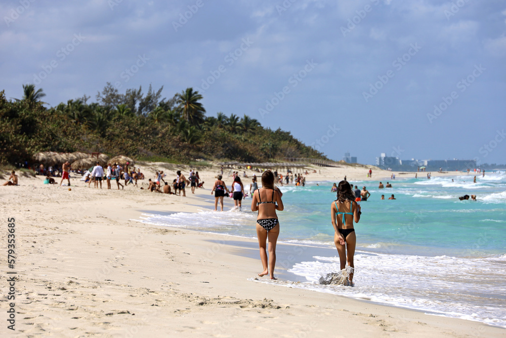 Two girls in bikini walking by the sand on tropical beach on swimming people and coconut palm trees background. Ocean coast, tourist resort in Varadero, Cuba