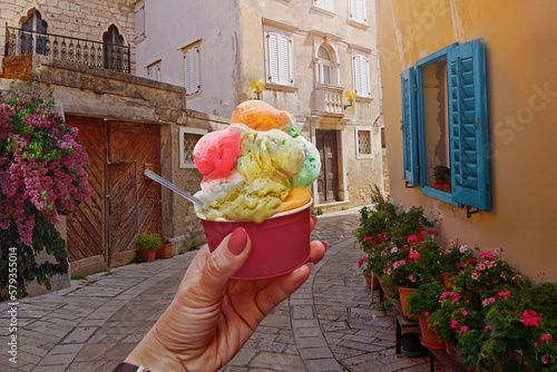 Photography Woman hand hold  beautiful bright sweet ice - cream cone with different flavors  held in hand on the background of old street  in  Porec,Croatia
