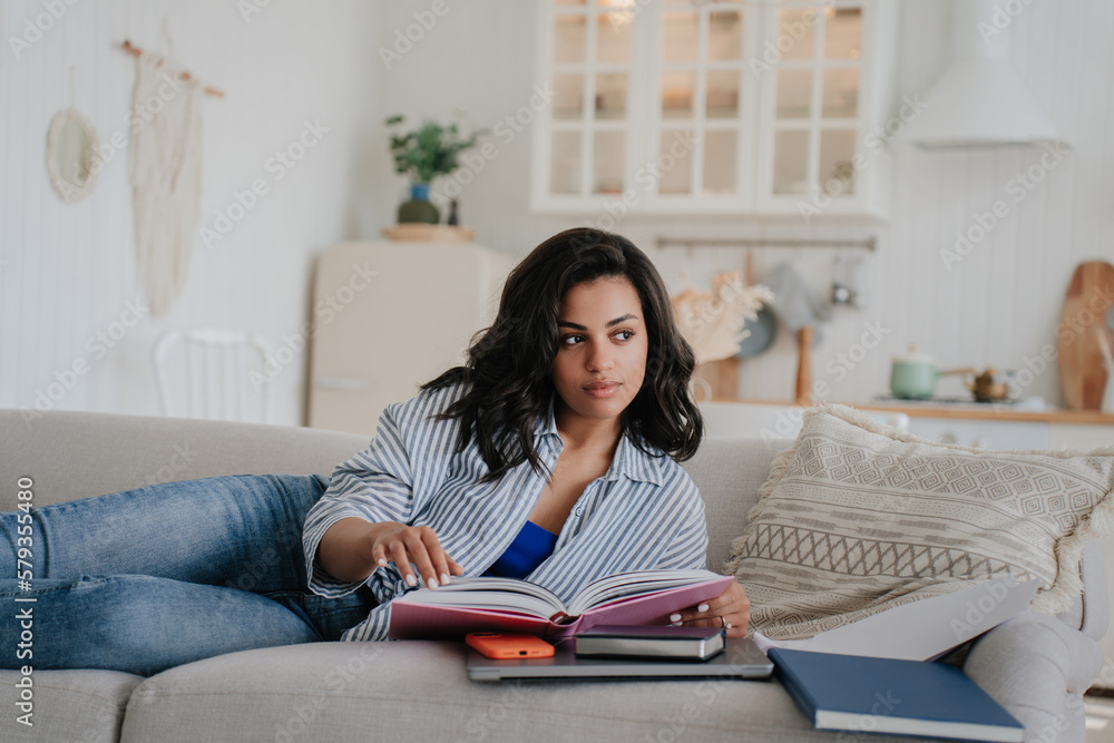 Thoughtful African American girl in casual laying on cozy couch reads book having break of remote work. Brazilian businesswoman explores catalogue of furniture. Young woman resting on vacation. Thoughtful African American girl in casual laying on cozy couch reads book having break of remote work. Brazilian businesswoman explores catalogue of furniture. Young woman resting on vacation.