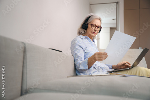 Concentrated gray-haired woman reads a summary at home on couch