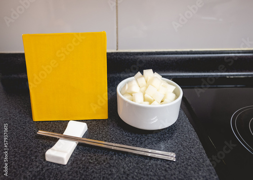 Traditional turnip kimchi in white bowl, metal chopstick and yellow board with copy space in table in kitchen (kimchi: pickled and fermented vegetables, garlic, salt, ginger, etc)