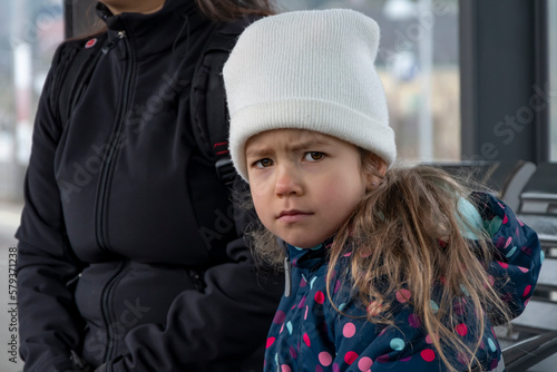 A little girl in a hat is sitting at a glassed-in bus stop with her mother waiting for public transport, a dissatisfied look on her face, an emotional portrait.