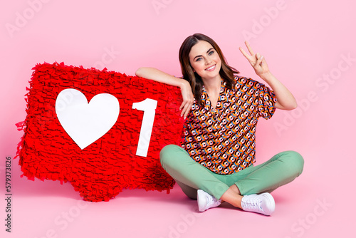 Full body size photo of young cheerful girl sitting floor show v-sign popular blogger pinata like huge symbol isolated on pink color background