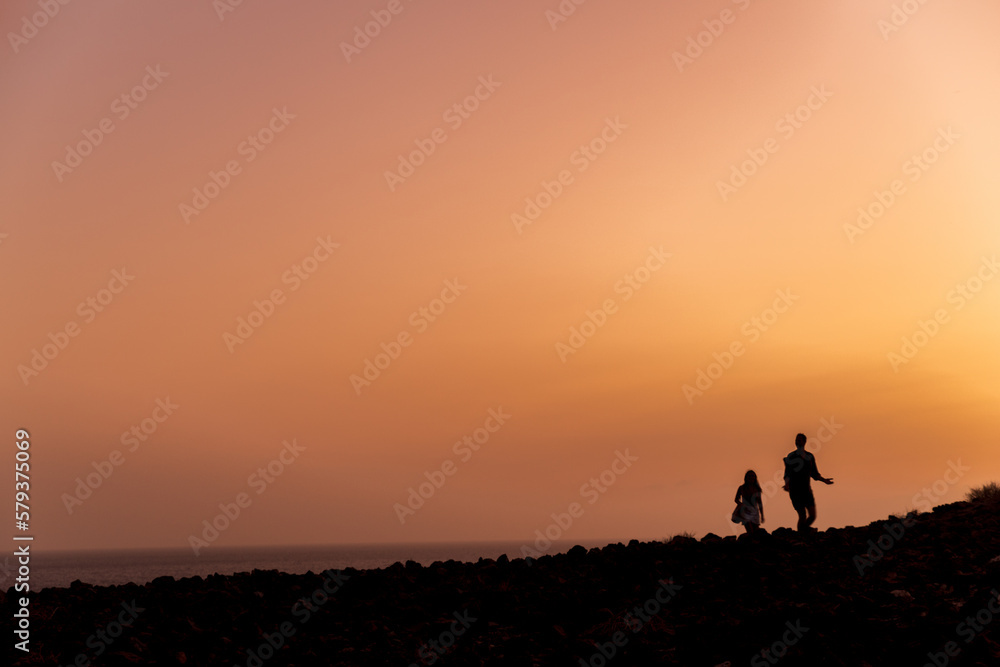 Silhouettes of a couple, tall man and short man, walking across rocky landscape with a deep red sunset on Tenerife Canary Islands