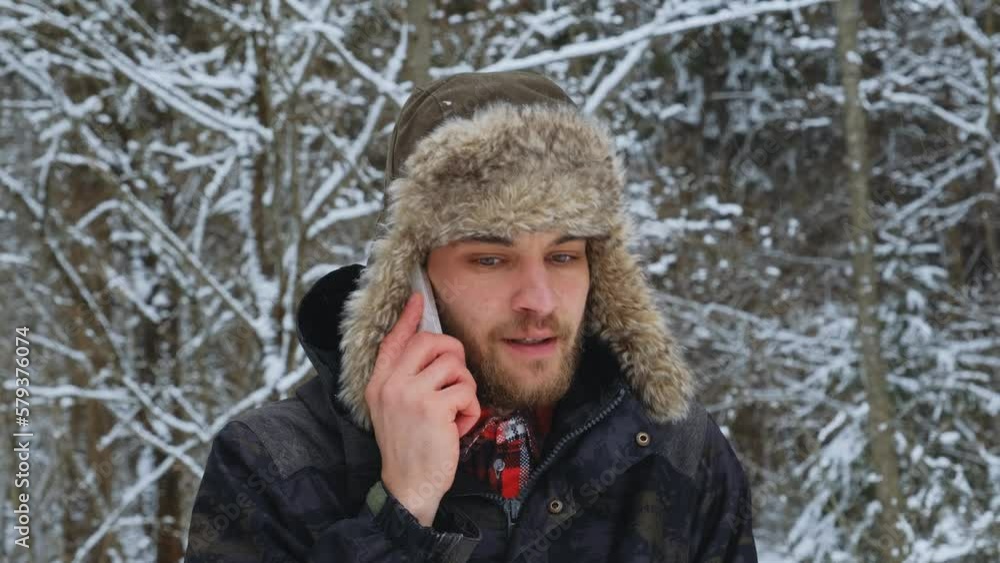 A young Caucasian man on a walk outside in winter wears a fluffy hat with earflaps and talks on the phone. The guy is frozen in the forest and is trying to keep warm by moving from side to side. A young Caucasian man on a walk outside in winter wears a fluffy hat with earflaps and talks on the phone. The guy is frozen in the forest and is trying to keep warm by moving from side to side.