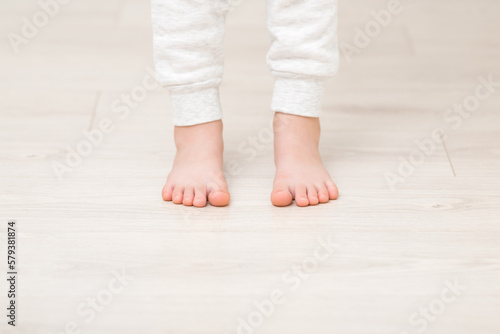 Little child legs in white warm trousers standing on light beige wooden floor at home room. Barefoot closeup. Front view.