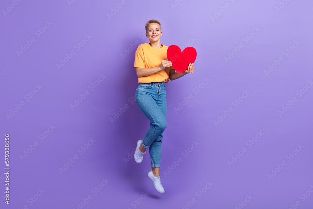 Full length photo of cheerful pleasant girl dressed jeans white sneakers jumping hold red paper heart isolated on violet color background Full length photo of cheerful pleasant girl dressed jeans white sneakers jumping hold red paper heart isolated on violet color background